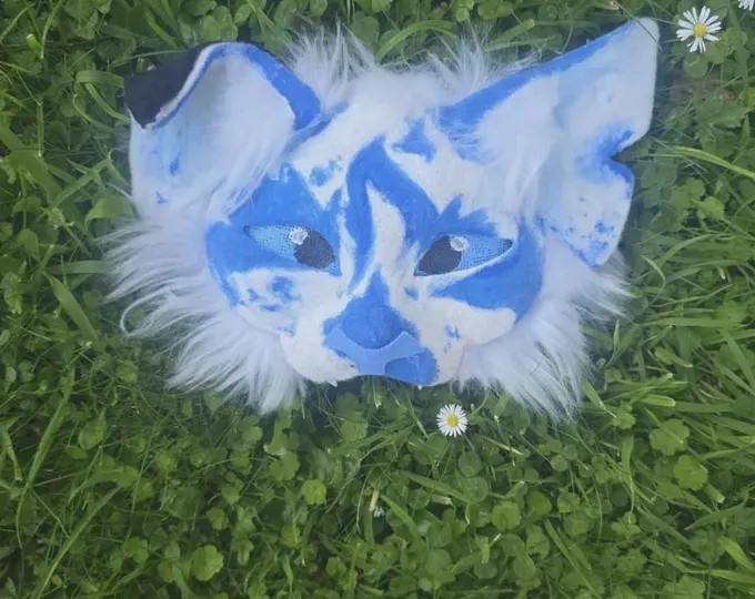 Blue and white patterned therian mask photographed in grass with daisies
