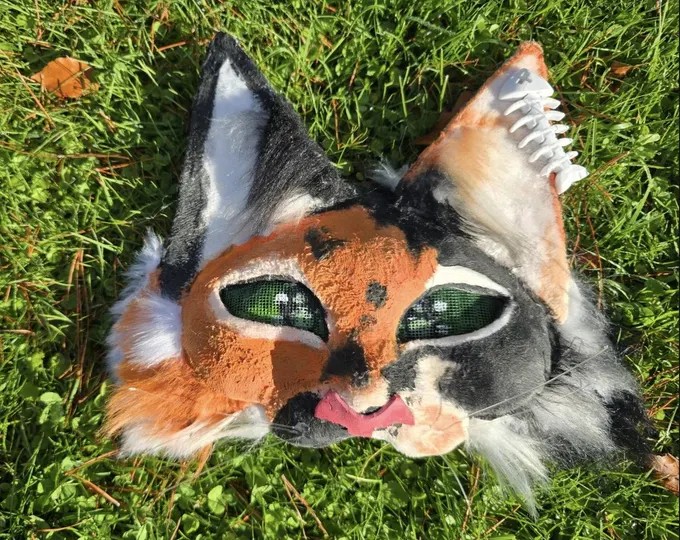Calico therian mask with green mesh eyes and pink tongue, photographed in grass
