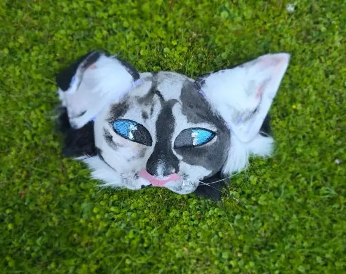 Grey and white therian mask with blue mesh eyes, photographed in grass