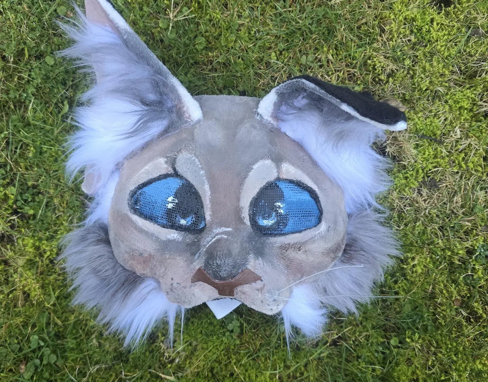 Hand-sculpted lynx therian mask with pale grey-tan faux fur and mesh eyes, photographed in the grass
